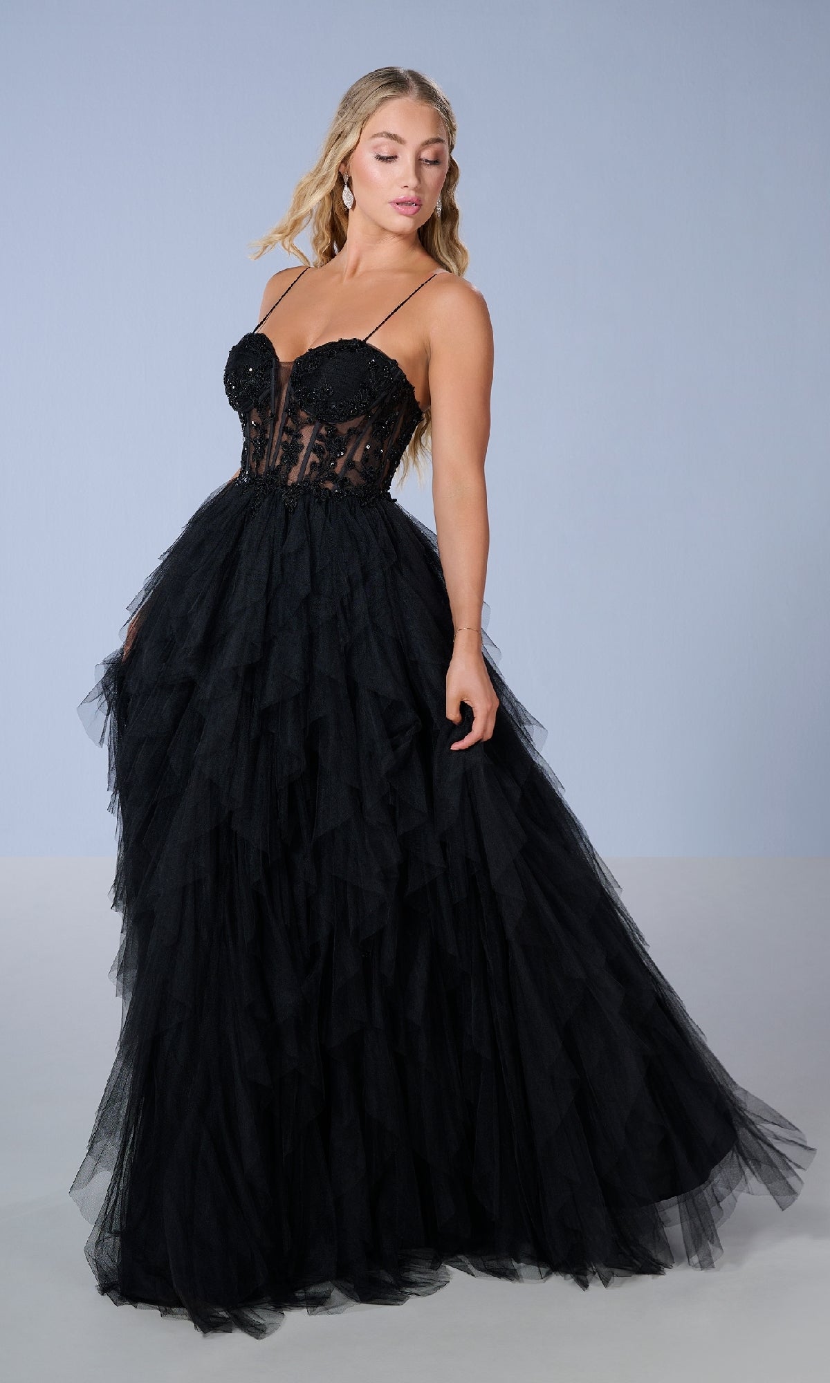 A woman stands against a plain light background wearing the Tiffany 16149 long prom dress, a black strapless ball gown with a layered tulle skirt and fitted semi-sheer bodice.